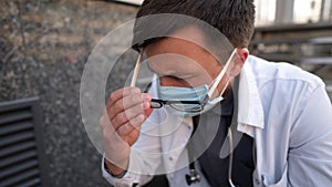 Caucasian young doctor man sits down on the stairs near the clinic building, tired and unhappy rubbing his nose and eyes