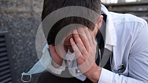 Caucasian young doctor man sits down on the stairs near the clinic building, tired and unhappy rubbing his nose and eyes