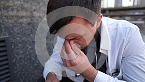 Caucasian young doctor man sits down on the stairs near the clinic building, tired and unhappy rubbing his nose and eyes