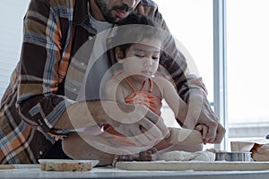 Caucasian young father with beard holding his little daughter hands to knead dough with rolling pin in kitchen