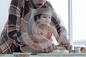 Caucasian young father with beard holding his little daughter hands to knead dough with rolling pin in kitchen