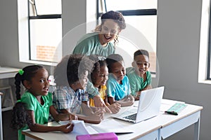 Caucasian young female teacher looking at african american elementary students while teaching laptop