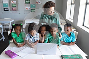 Caucasian young female teacher standing by african american elementary students using laptop at desk