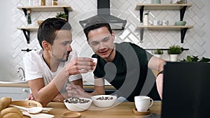 Caucasian young gay couple is sitting at the kitchen table eating breakfast and watching movie in the morning at kitchen