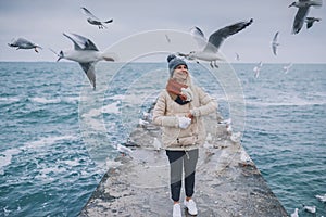 Caucasian young happy woman feeds seagulls on the sea. Pretty female smiling and wearing coat, scarf and watching flying seagulls