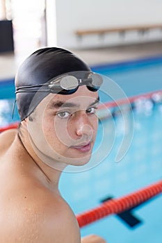 Caucasian young male swimmer standing by pool indoors, looking at camera