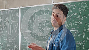 Caucasian young male teacher standing at blackboard with formulas, graphics and laws and explaining with finger