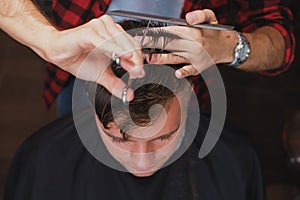 Caucasian young man in Barbershop. Barber is cuttinhg his hair in hipster style.