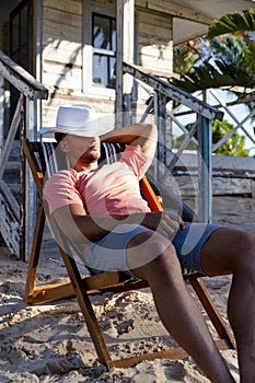Caucasian young man with hat on face sitting on deck chair outside cottage at beach