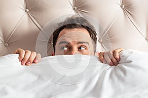 Caucasian young man hiding in bed under the blanket at home