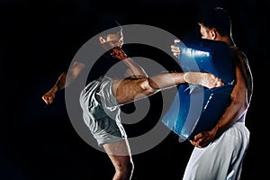 Caucasian young man holding a punching bag while his friend kickikng punches at it