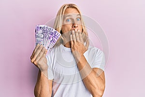 Caucasian young man with long hair holding 100 philippine peso banknotes covering mouth with hand, shocked and afraid for mistake