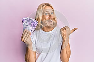 Caucasian young man with long hair holding 100 philippine peso banknotes pointing thumb up to the side smiling happy with open