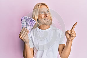 Caucasian young man with long hair holding 100 philippine peso banknotes smiling happy pointing with hand and finger to the side