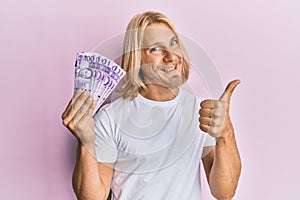 Caucasian young man with long hair holding 100 philippine peso banknotes smiling happy and positive, thumb up doing excellent and
