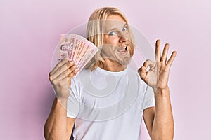 Caucasian young man with long hair holding 100 thai baht banknotes doing ok sign with fingers, smiling friendly gesturing