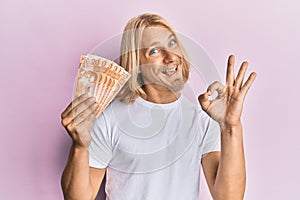 Caucasian young man with long hair holding 20 philippine peso banknotes doing ok sign with fingers, smiling friendly gesturing