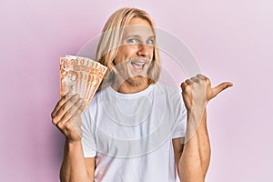 Caucasian young man with long hair holding 20 philippine peso banknotes pointing thumb up to the side smiling happy with open