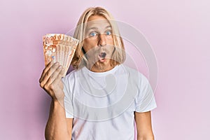 Caucasian young man with long hair holding 20 philippine peso banknotes scared and amazed with open mouth for surprise, disbelief