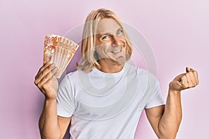 Caucasian young man with long hair holding 20 philippine peso banknotes screaming proud, celebrating victory and success very