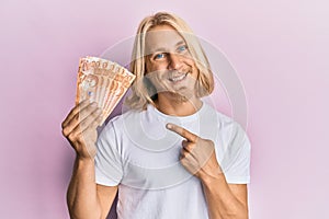 Caucasian young man with long hair holding 20 philippine peso banknotes smiling happy pointing with hand and finger