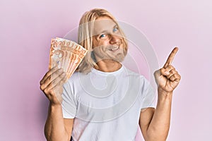 Caucasian young man with long hair holding 20 philippine peso banknotes smiling happy pointing with hand and finger to the side