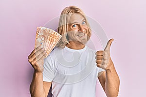 Caucasian young man with long hair holding 20 philippine peso banknotes smiling happy and positive, thumb up doing excellent and