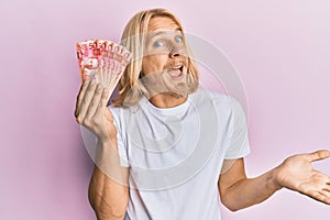 Caucasian young man with long hair holding 50 philippine peso banknotes celebrating achievement with happy smile and winner