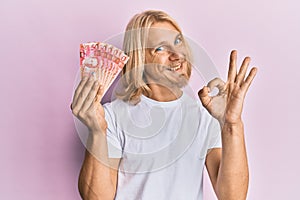 Caucasian young man with long hair holding 50 philippine peso banknotes doing ok sign with fingers, smiling friendly gesturing
