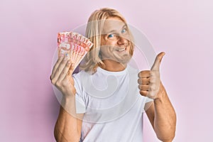 Caucasian young man with long hair holding 50 philippine peso banknotes smiling happy and positive, thumb up doing excellent and