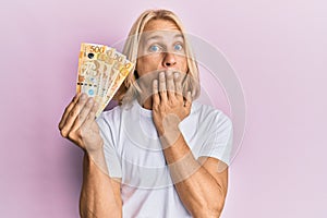 Caucasian young man with long hair holding 500 philippine peso banknotes covering mouth with hand, shocked and afraid for mistake