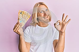 Caucasian young man with long hair holding 500 philippine peso banknotes doing ok sign with fingers, smiling friendly gesturing