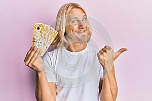 Caucasian young man with long hair holding 500 philippine peso banknotes pointing thumb up to the side smiling happy with open