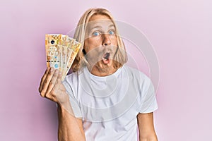 Caucasian young man with long hair holding 500 philippine peso banknotes scared and amazed with open mouth for surprise, disbelief
