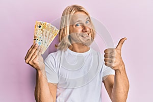 Caucasian young man with long hair holding 500 philippine peso banknotes smiling happy and positive, thumb up doing excellent and