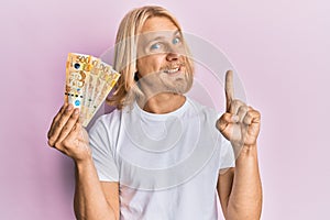 Caucasian young man with long hair holding 500 philippine peso banknotes smiling with an idea or question pointing finger with