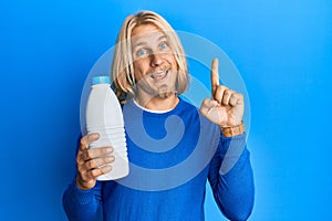 Caucasian young man with long hair holding liter bottle of milk smiling with an idea or question pointing finger with happy face,