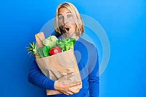 Caucasian young man with long hair holding paper bag with groceries scared and amazed with open mouth for surprise, disbelief face