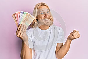 Caucasian young man with long hair holding philippine peso banknotes screaming proud, celebrating victory and success very excited