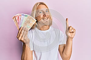 Caucasian young man with long hair holding philippine peso banknotes smiling with an idea or question pointing finger with happy