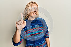 Caucasian young man with long hair wearing colorful winter sweater smiling with an idea or question pointing finger up with happy