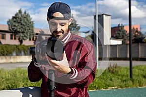 Caucasian young man with reverse cap and red jacket works outdoor with his camera on a gimbal stabiliser. Video operator focusing