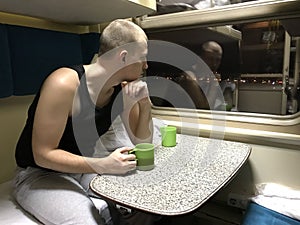 Caucasian young man sits at table in train car, holds green cup of tea and looks thoughtfully out window at lights of night city