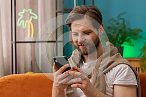 Caucasian young man sitting on sofa, using smartphone share messages on social media application