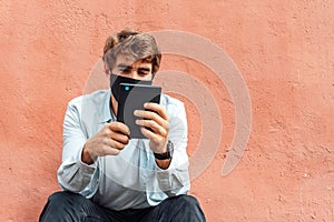 Caucasian young man wearing a mask and reading and ebook