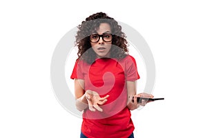 caucasian young pretty curly brunette woman in a red t-shirt spreads her arms in excitement holding a smartphone in her