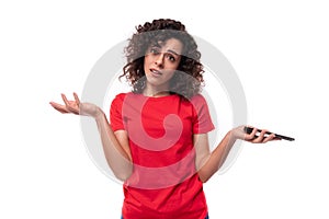 caucasian young pretty curly brunette woman in a red t-shirt spreads her arms in excitement holding a smartphone in her