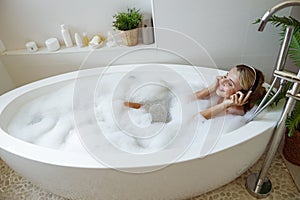 Caucasian young pretty woman in headphones lying in bathtub with foam and listening to music