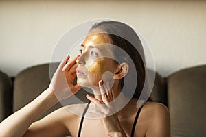 Caucasian young woman applying moisturizing or peeling golden facial mask and touch her face with hands. Side view.