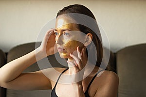 Caucasian young woman applying moisturizing or peeling golden facial mask and touch her face with hands. Side view.